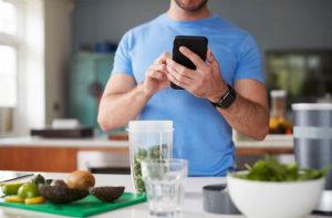 A man looking at a smartphone. Before him is diced produce, some in a blender cup